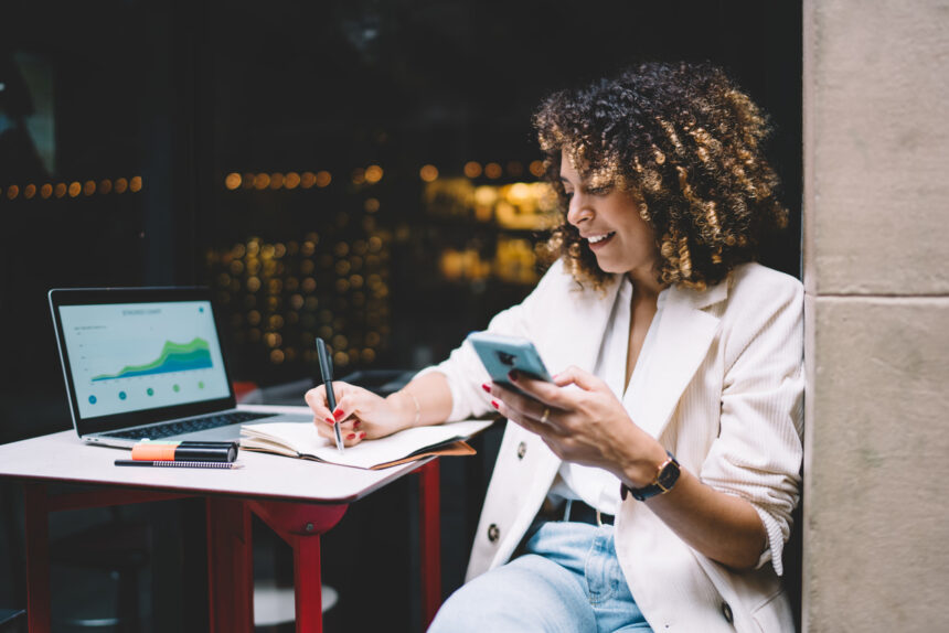 Happy female IT professional working remotely with web projecting and online diagrams displayed on laptop screen, cheerful woman with smartphone device in hand writing informative notes in textbook