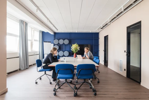 Two colleagues talking across a white table with laptops in a meeting room with a navy blue feature wall.