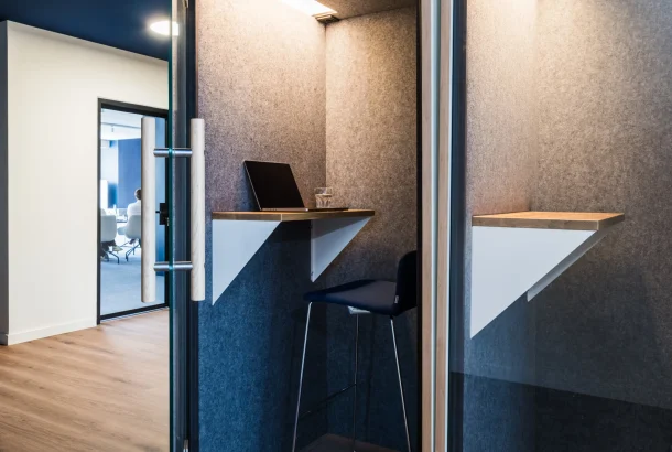 Glass-doored phone booths with felt-lined walls, a wall-mounted wooden standing desk, laptop and a navy bar stool.